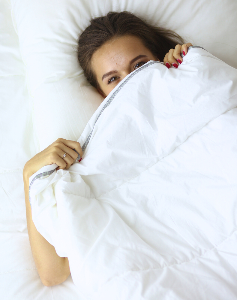 A pretty young woman peeking from under the covers in her bedroom happilly.